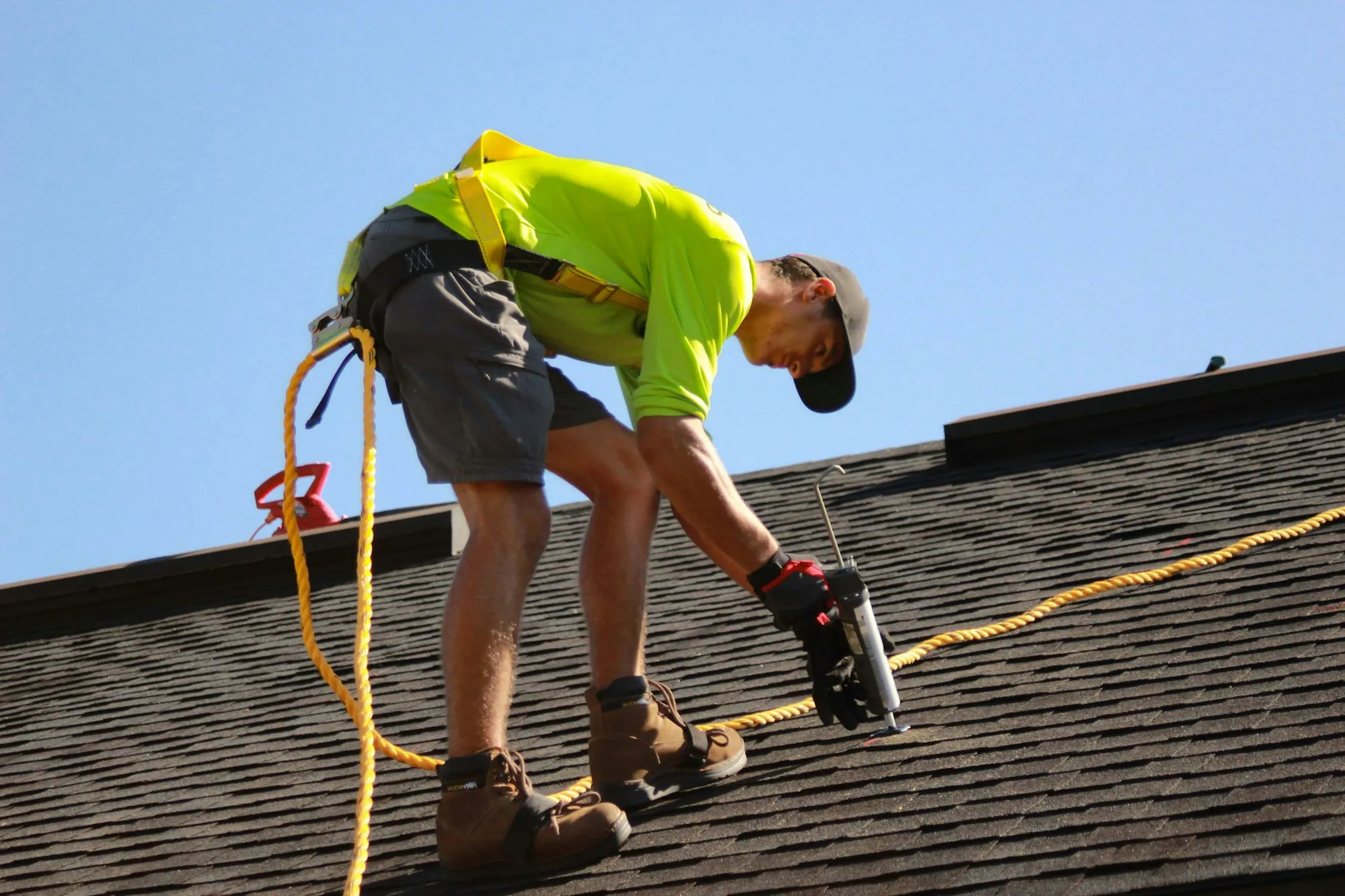 Roofer in a safety harness using a caulk gun on a shingle roof