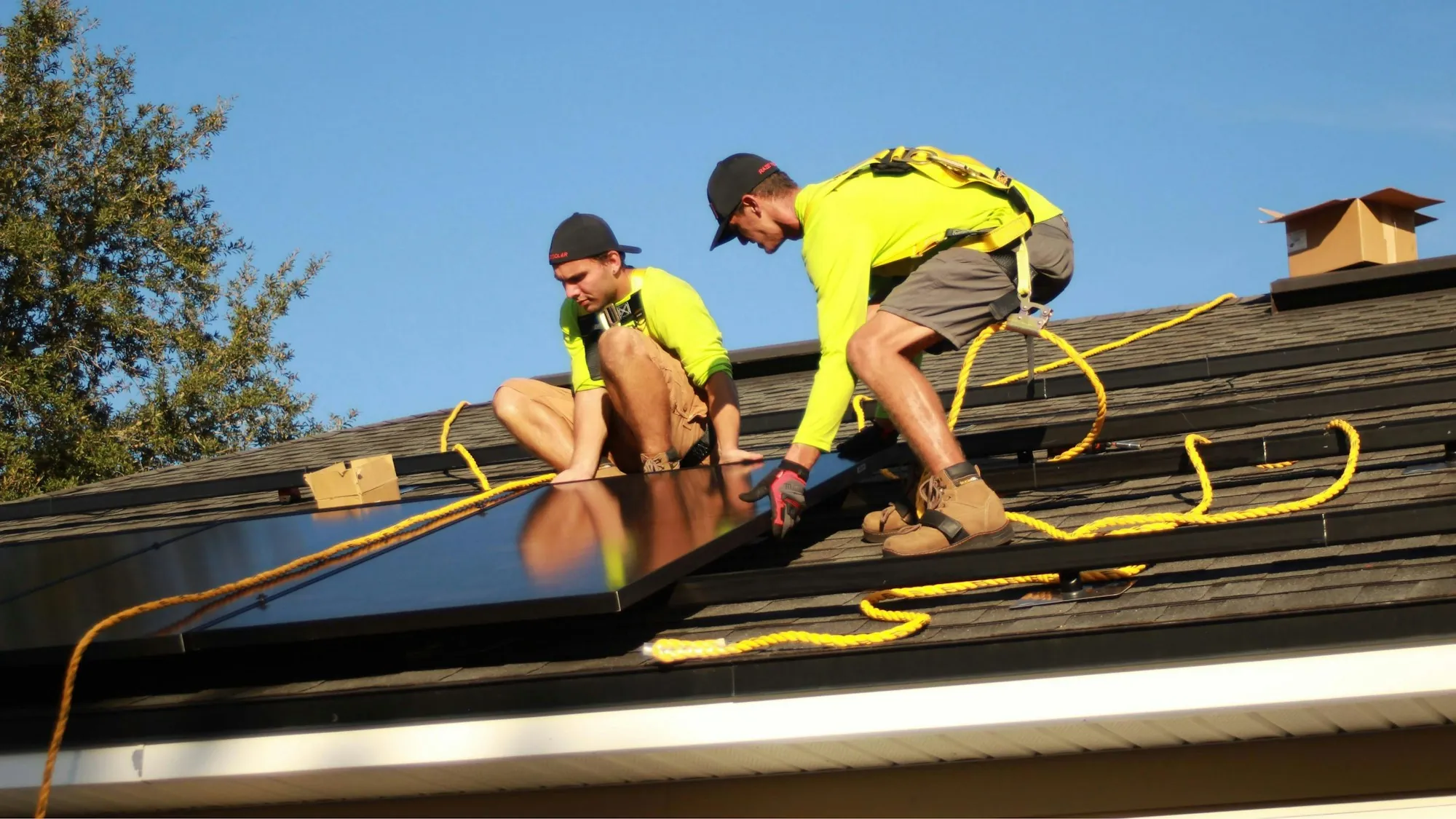 Two installers kneeling on a shingle roof while positioning a solar panel